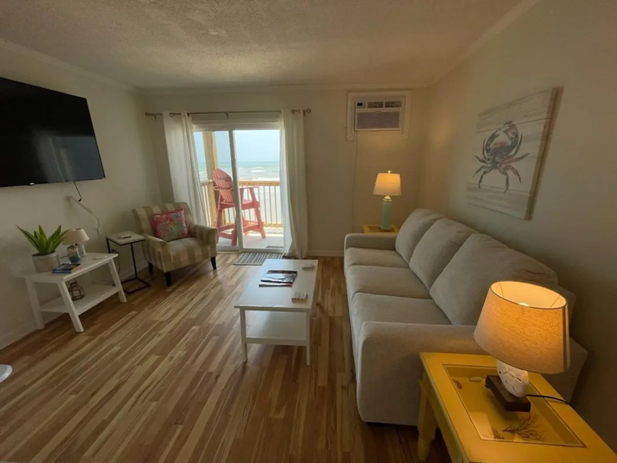 This image shows a cozy living room with a comfortable sofa positioned to enjoy both the TV and the ocean view through large windows. Chairs on the private balcony are visible just outside, inviting relaxation and connection with the outdoors. Warm lighting and tasteful décor create an inviting atmosphere for unwinding or enjoying a movie night.