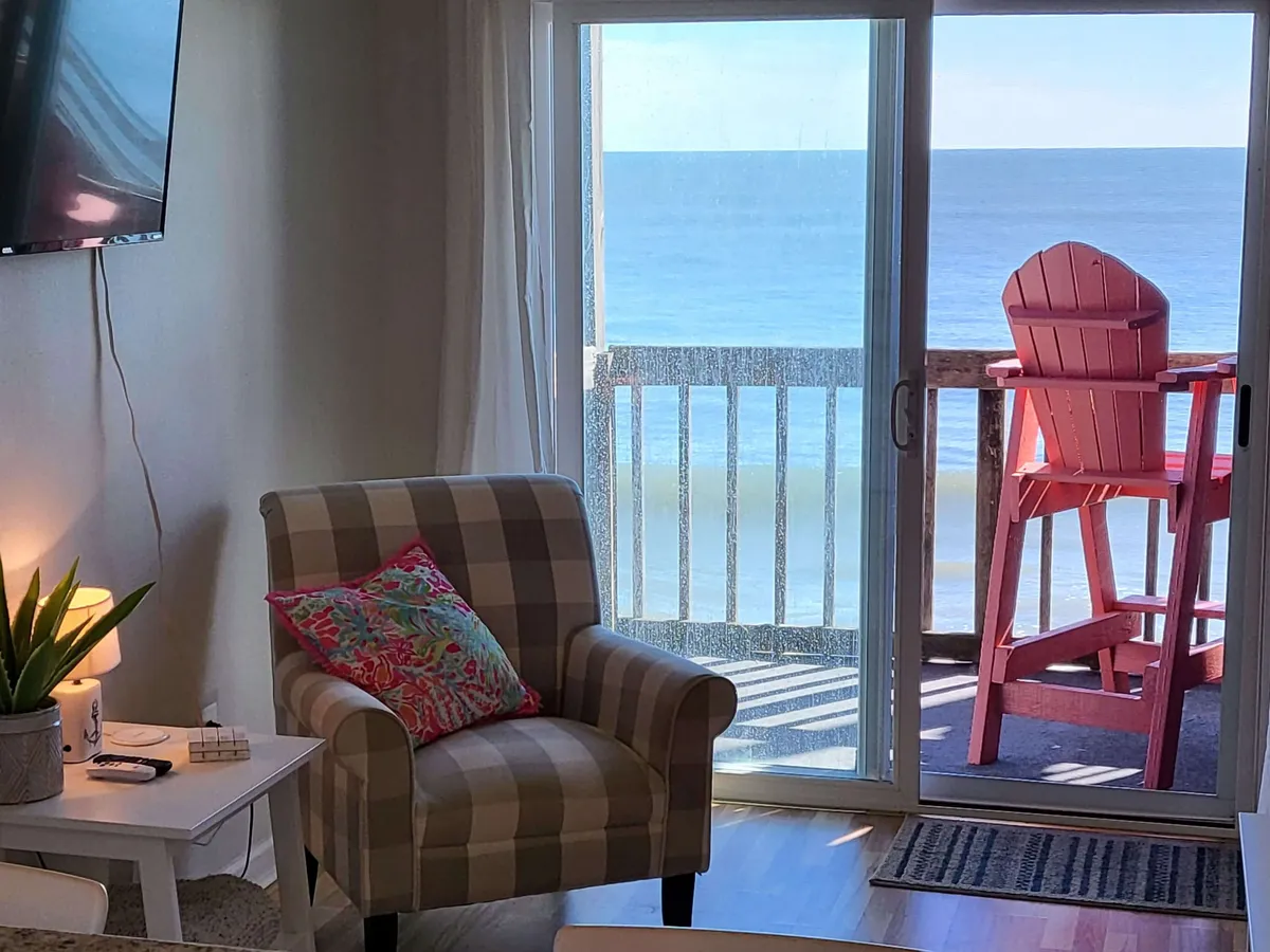 This interior view features a checkerboard armchair with a colorful throw pillow facing a sliding glass door that opens to a private balcony furnished with a red Adirondack chair. Through the door, a stunning ocean view awaits, capturing the essence of a breezy coastal retreat.