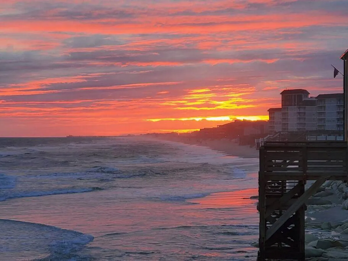 A tranquil sunset scene featuring the ocean, a pier stretching into the water, and soft clouds overhead. People can be seen walking along the beach, fishing, or simply watching the sun dip below the horizon, encapsulating the peaceful end to a perfect day on the coast.