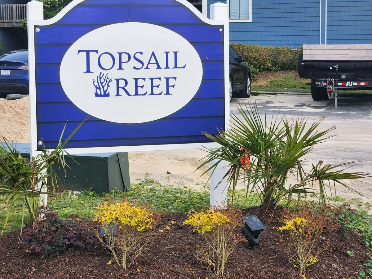 This image shows the Topsail Reef property sign welcoming guests, with manicured landscaping and vehicles parked nearby. The inviting entrance is set against a backdrop of open sky and hints at the property's proximity to local beaches and dining.