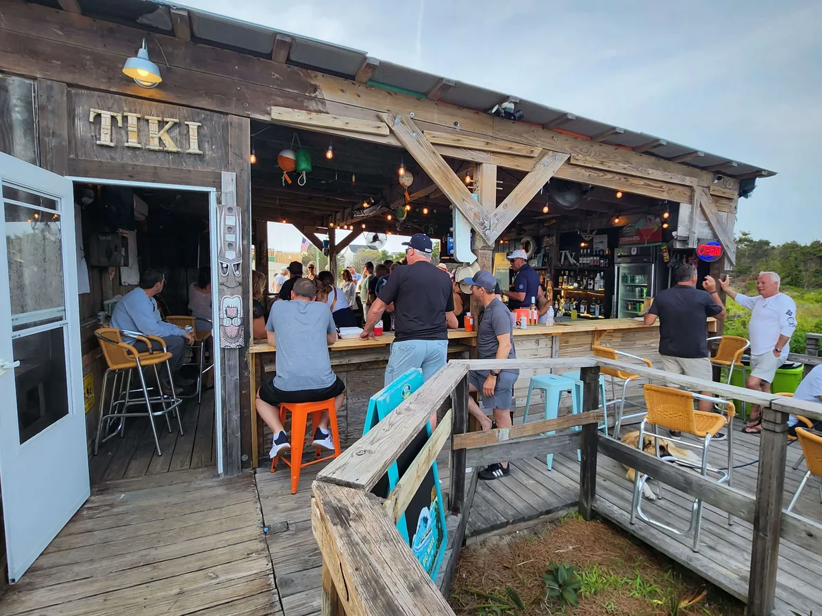 A lively outdoor Tiki bar scene, with a crowd gathered around colorful seating and an open doorway. The festive mood is enhanced by people socializing, sipping drinks, and enjoying the vibrant local atmosphere, suggesting a blend of relaxation and local flavor.