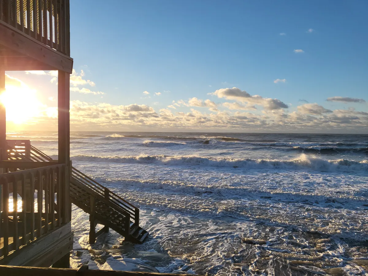 A tranquil sunset washes over the beach, with gentle waves and a wooden structure leading down to the sand. The soft light creates a peaceful ambiance, ideal for winding down together after a day of adventure.