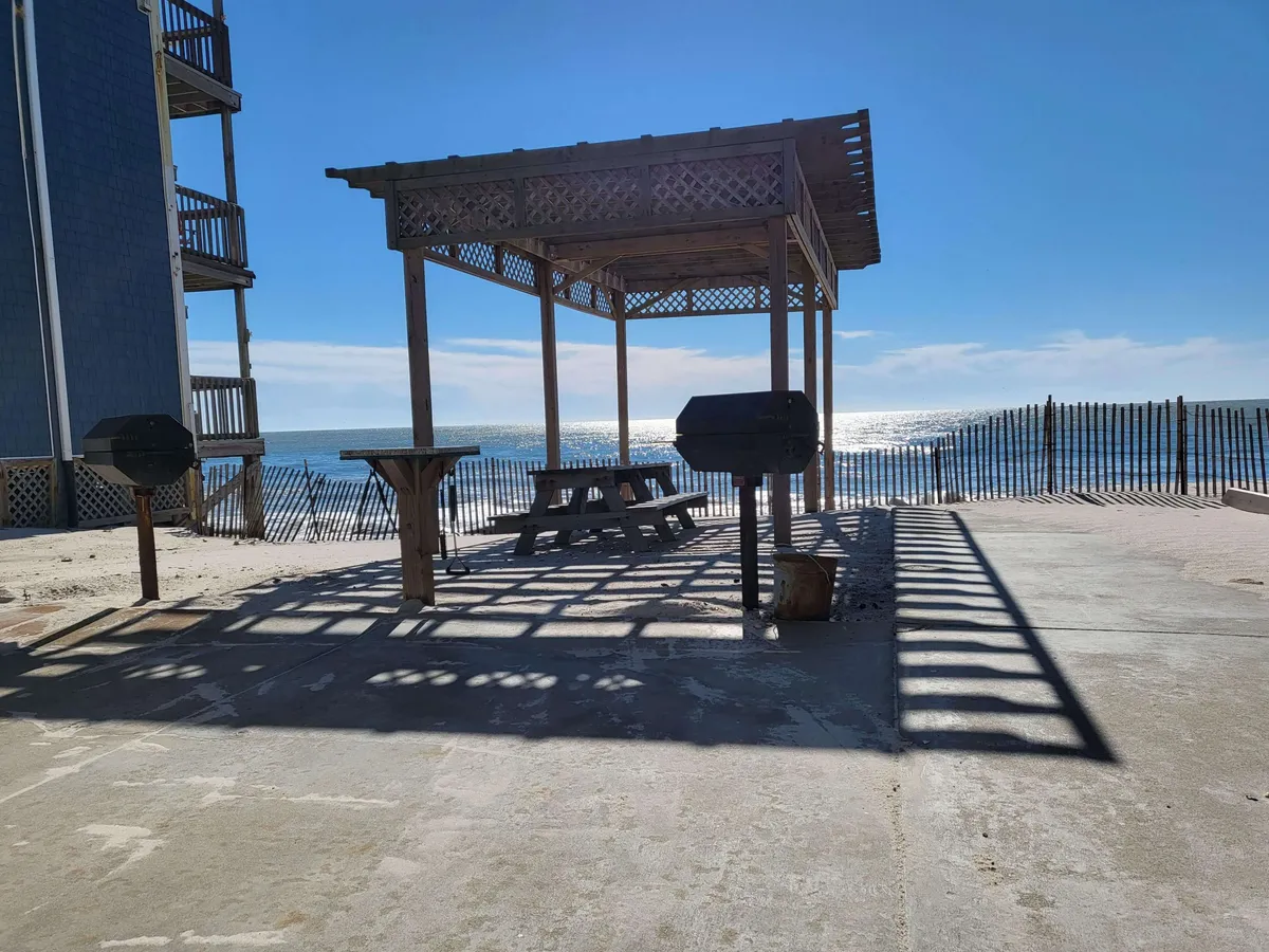 A welcoming wooden gazebo equipped with a picnic table and barbecue grills is set against a picturesque ocean view. The area feels lively and perfect for family gatherings or casual meals, with ample space for both adults and children to relax and socialize by the shore.