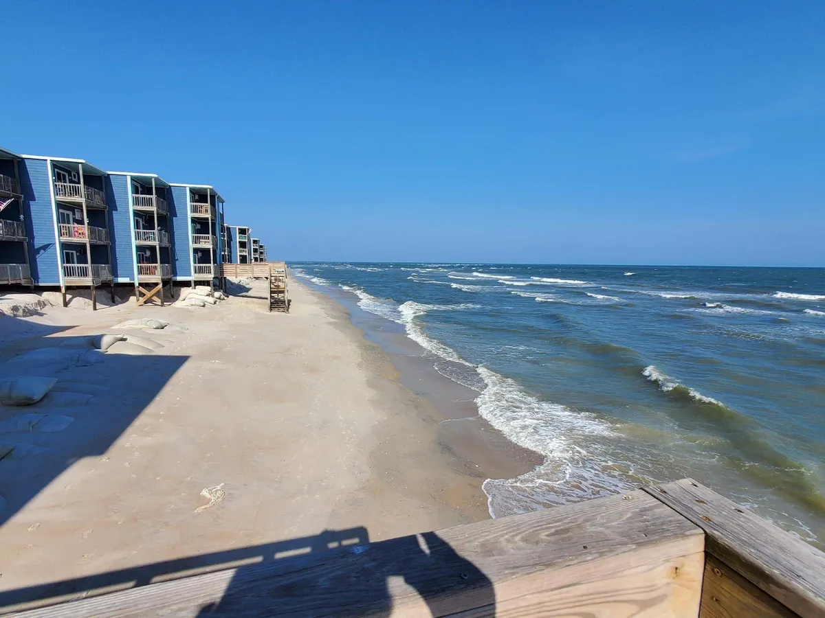 A wide view of the sandy beachfront with gentle ocean waves and several condominium buildings in the background. The atmosphere is calm and inviting, perfect for a day of sunbathing, beachcombing, or surfing. The scene conveys the immediate access to the coast and the laid-back vibe of North Topsail.