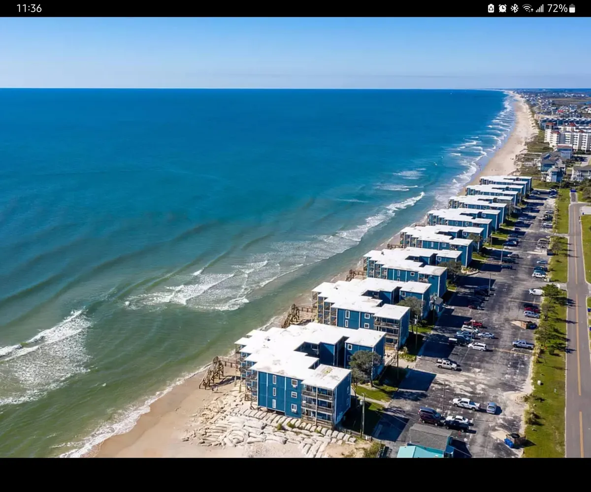 An expansive view of beachfront properties facing the rolling waves, with easy beach access and a visible parking area for convenience. The scene is calm and inviting, epitomizing serene mornings, beach walks, and proximity to local seafood dining options.