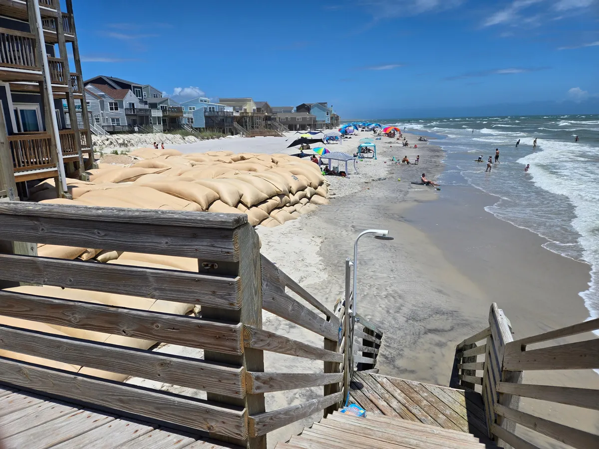 A lively beachfront scene featuring sandbags lining the shore, colorful beach umbrellas scattered across the sand, and waves crashing just steps from where guests sunbathe and play. Groups are engaged in various activities such as swimming, volleyball, and picnicking, with the ocean backdrop creating an energetic, vacation-like atmosphere.