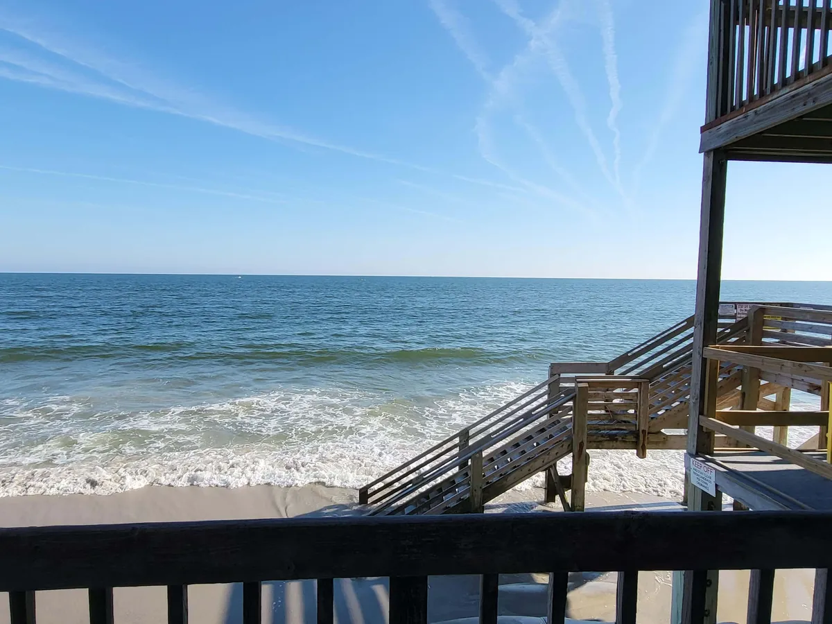 A relaxed exterior view showcasing a wooden staircase leading down to soft sand, with gentle ocean waves rolling in beneath a clear blue sky. The setting is tranquil, perfect for morning walks, yoga, and direct access from the property to the beach.