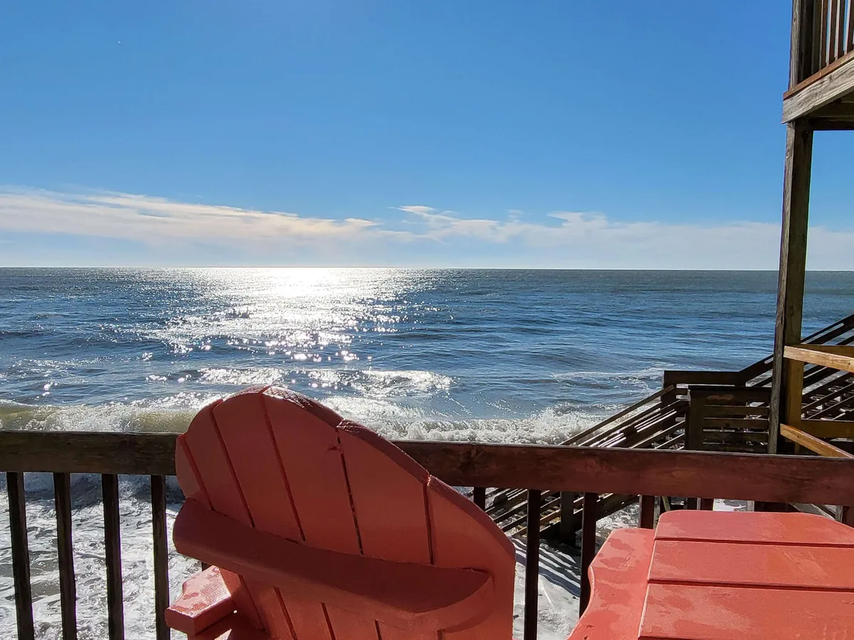 The image showcases a bright red Adirondack chair positioned on a private balcony, overlooking shimmering ocean waves as sunlight dances across the water. The scene is tranquil, with the promise of peaceful mornings spent watching the sunrise and unobstructed views of the beach just steps away. The inviting setup suggests moments of relaxation, reading, or quiet contemplation while being immersed in the soothing sounds and sights of the coast.