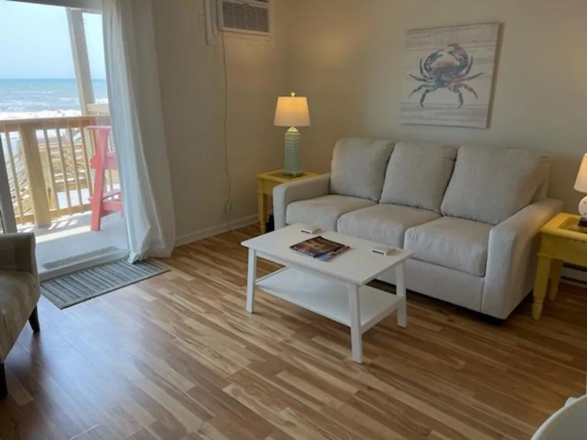 An inviting living area with a comfortable sofa, coffee table, and cheerful crab artwork. Large windows offer a glimpse of the ocean, and the space is filled with natural light, creating a welcoming environment for relaxation and social moments.