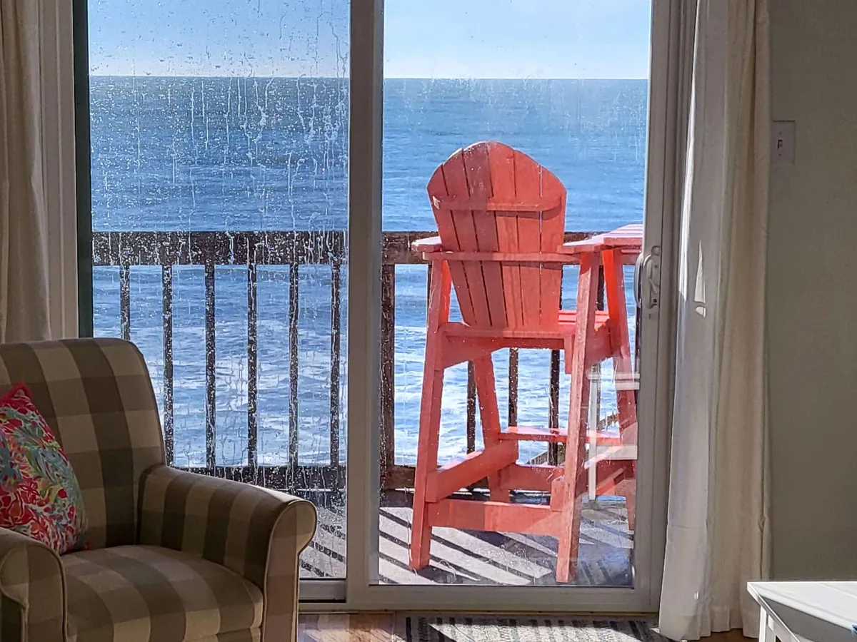 An inviting interior scene where a red high chair and a checkered armchair are positioned near light, airy curtains, framing a panoramic ocean view. The tranquil mood is ideal for savoring a morning coffee, reading, or quietly watching the sunset from the comfort of the living space.